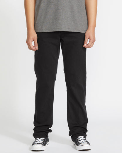 A person is seen from the waist down, wearing a gray t-shirt, Volcom Men's Solver Denim Pant in black, and black-and-white sneakers against a plain white background.