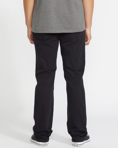 A person faces away from the camera, wearing a gray t-shirt, Volcom Men's Solver Denim Pant in black, and black-and-white sneakers, posed against a plain white background.