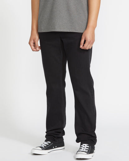 A person wears a gray t-shirt, black Volcom Men's Solver Denim Pant, and black low-top sneakers with white soles, standing against a plain white background. The image is cropped below the shoulders.