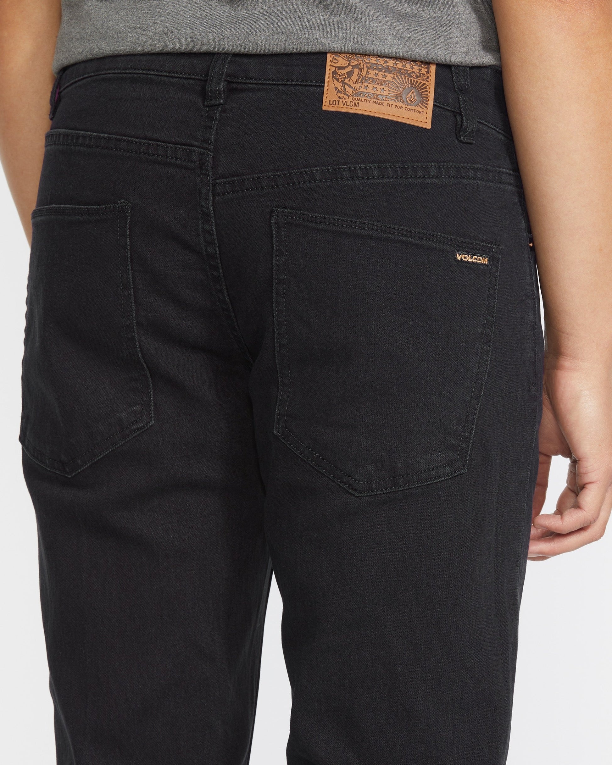A person is shown from the waist down and back wearing Volcom Men's Solver Denim Pant in black with back pockets, a brown leather patch on the waistband, and a small “Volcom” tag above the right pocket, paired with a gray shirt.