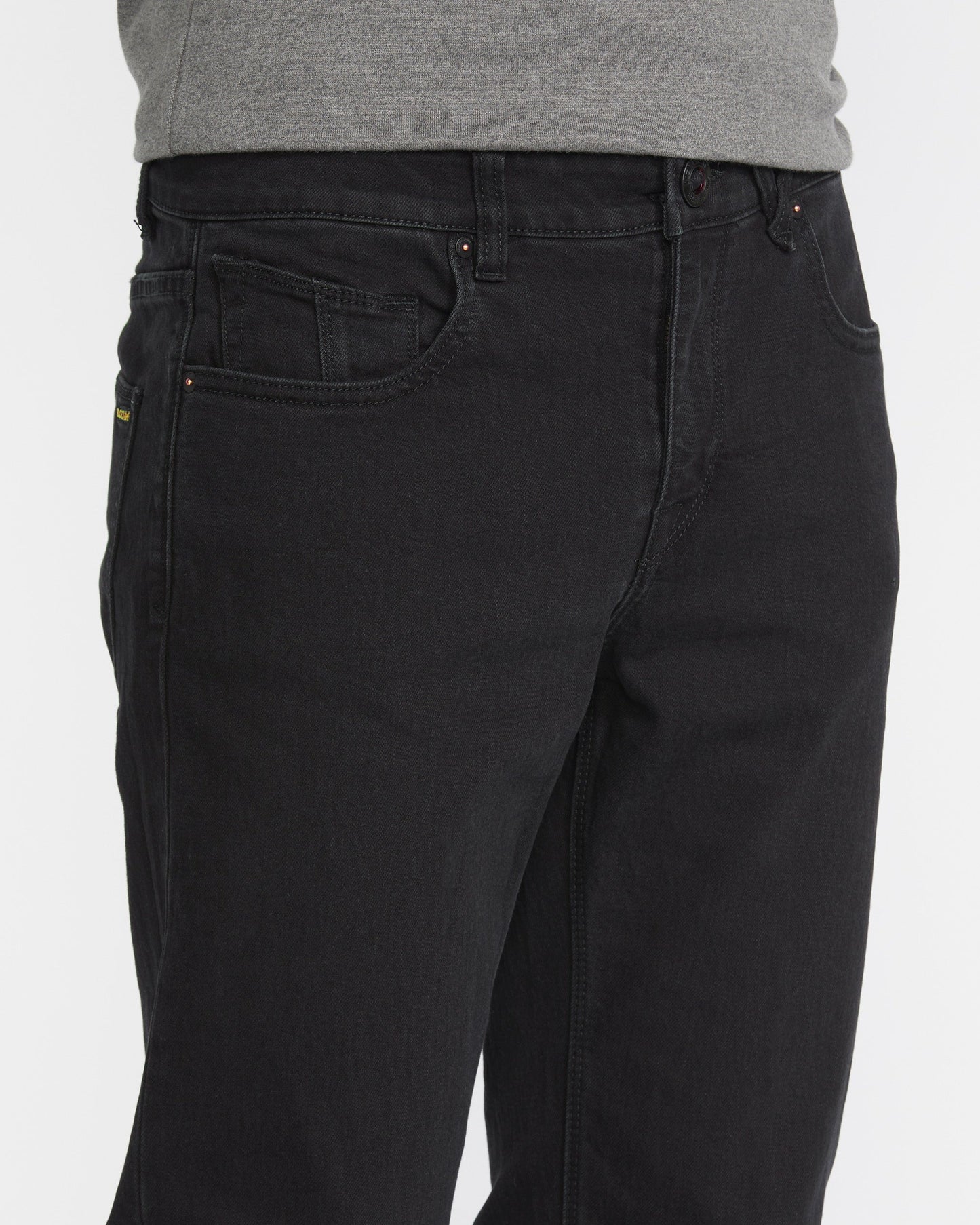 A man wears Volcom Men's Solver Denim Pant in black and a gray shirt, shown from waist to mid-thigh against a plain white background.