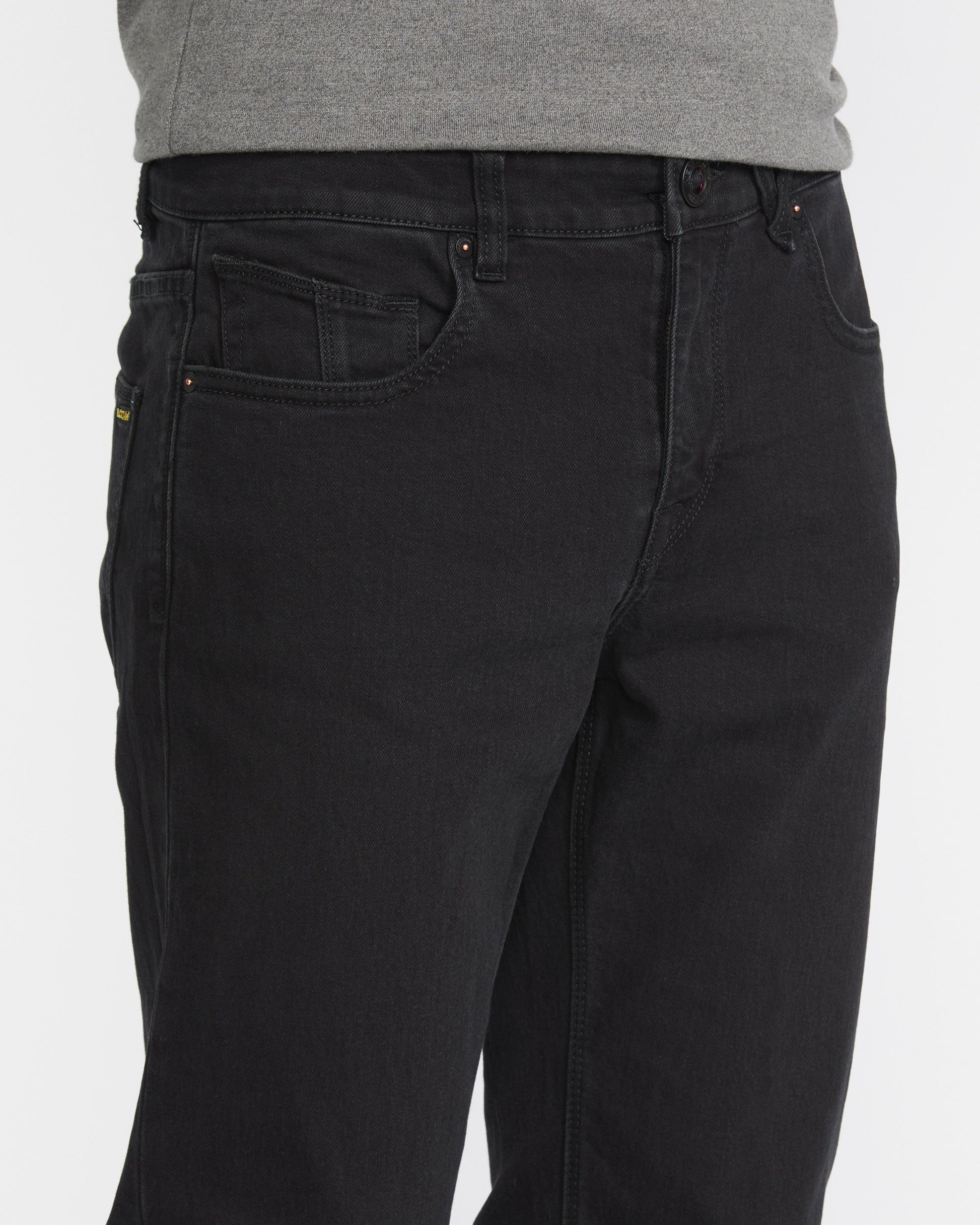 A man wears Volcom Men's Solver Denim Pant in black and a gray shirt, shown from waist to mid-thigh against a plain white background.