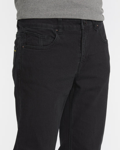A man wears Volcom Men's Solver Denim Pant in black and a gray shirt, shown from waist to mid-thigh against a plain white background.