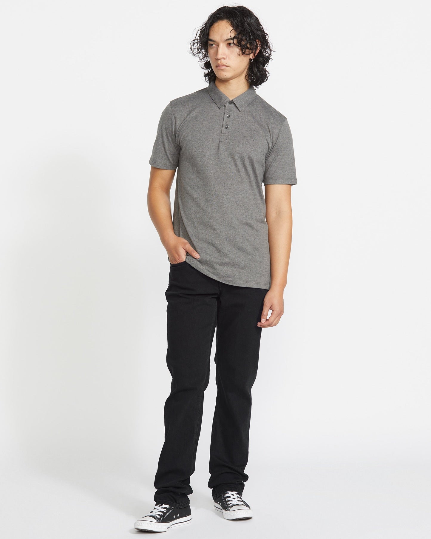 A person with medium-length curly hair, in a gray short-sleeve polo shirt, Volcom Men's Solver Denim Pant in black, and black-and-white sneakers stands against a plain white background, glancing slightly to the side.