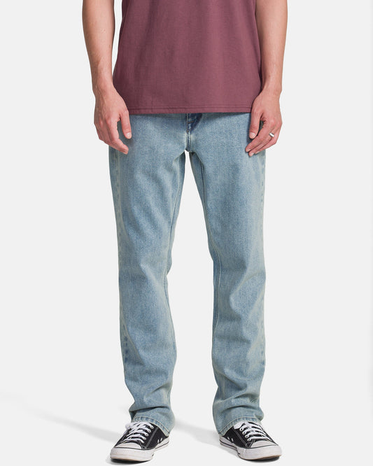 A person wearing a maroon t-shirt, Volcom Men's Solver Denim Pants in Dust Bowl Indigo, and black low-top sneakers stands against a plain white background. The image captures them from shoulders to feet.
