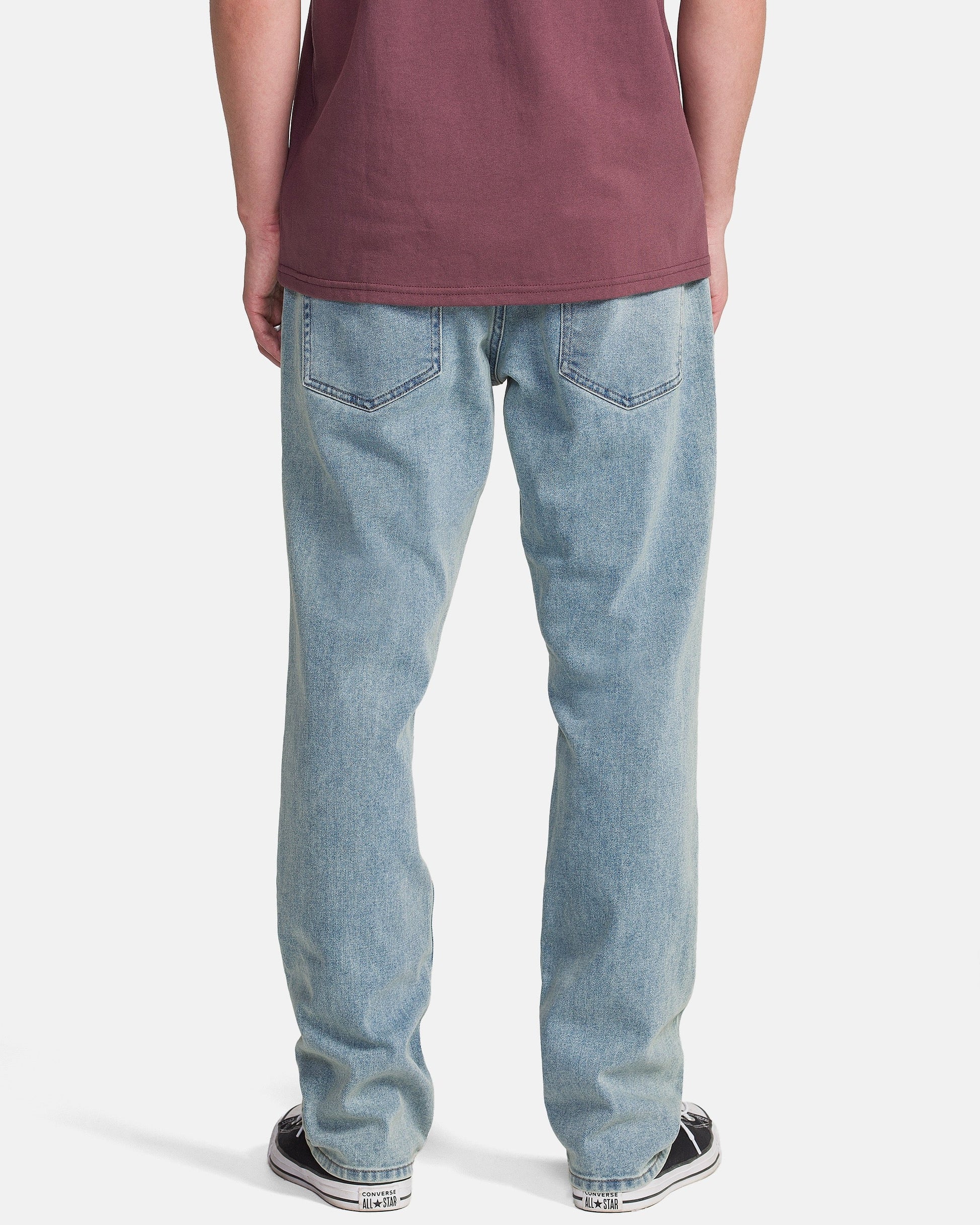 A person stands with their back to the camera, wearing Volcom Men's Solver Denim Pants in Dust Bowl Indigo, a maroon t-shirt, and black-and-white sneakers against a white background.
