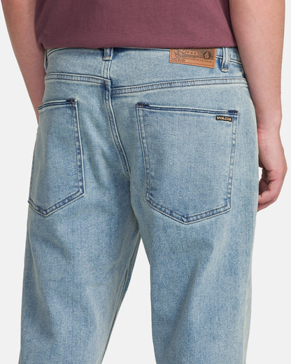 A person is shown from the back wearing Men's Solver Denim Pants by Volcom in Dust Bowl Indigo with a maroon shirt. The jeans feature two large back pockets and a brown Volcom patch on the waistband.