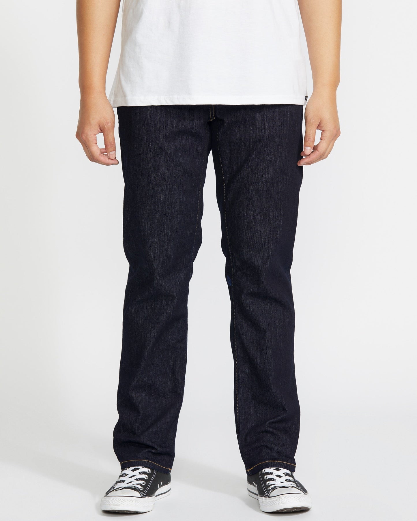 A person stands against a plain white background, wearing a white tee and Volcom Men's Solver Denim Pant in dark blue stretch denim. The image is cropped at the shoulders to focus on the jeans and black-and-white sneakers.