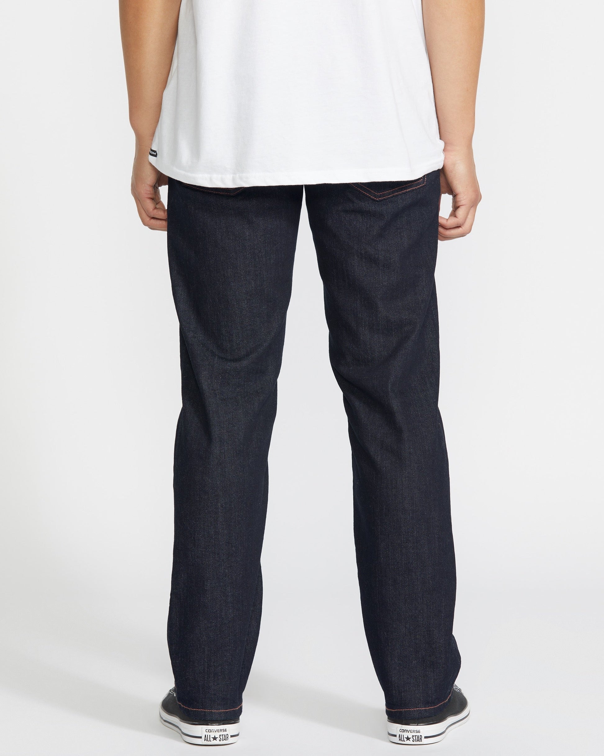 A person stands with their back to the camera, wearing a white T-shirt, white sneakers, and Volcom Men's Solver Denim Pant in dark blue stretch denim against a plain white background.