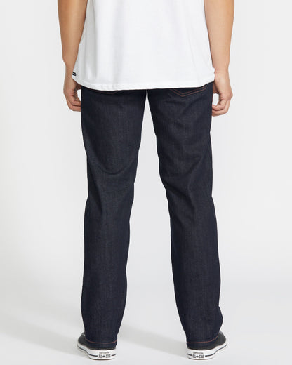 A person stands with their back to the camera, wearing a white T-shirt, white sneakers, and Volcom Men's Solver Denim Pant in dark blue stretch denim against a plain white background.