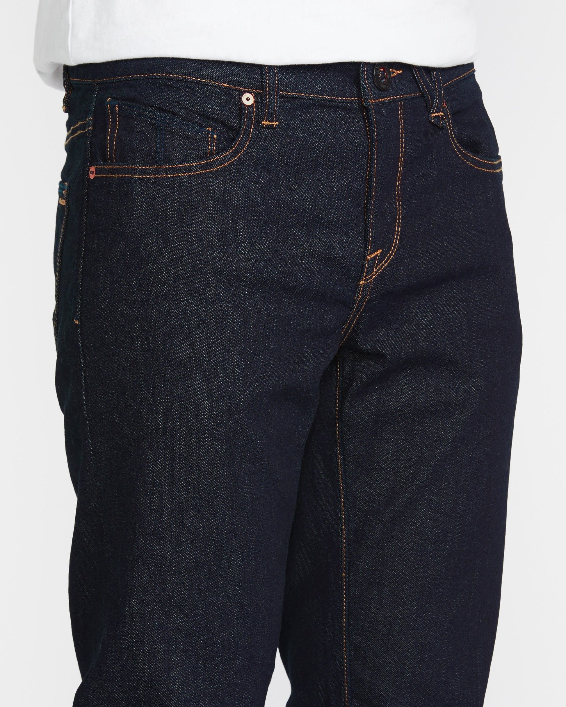 A close-up of a person wearing Volcom Men's Solver Denim Pant in dark blue stretch denim with modern straight leg, orange stitching, and a white shirt.
