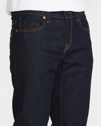 A close-up of a person wearing Volcom Men's Solver Denim Pant in dark blue stretch denim with modern straight leg, orange stitching, and a white shirt.