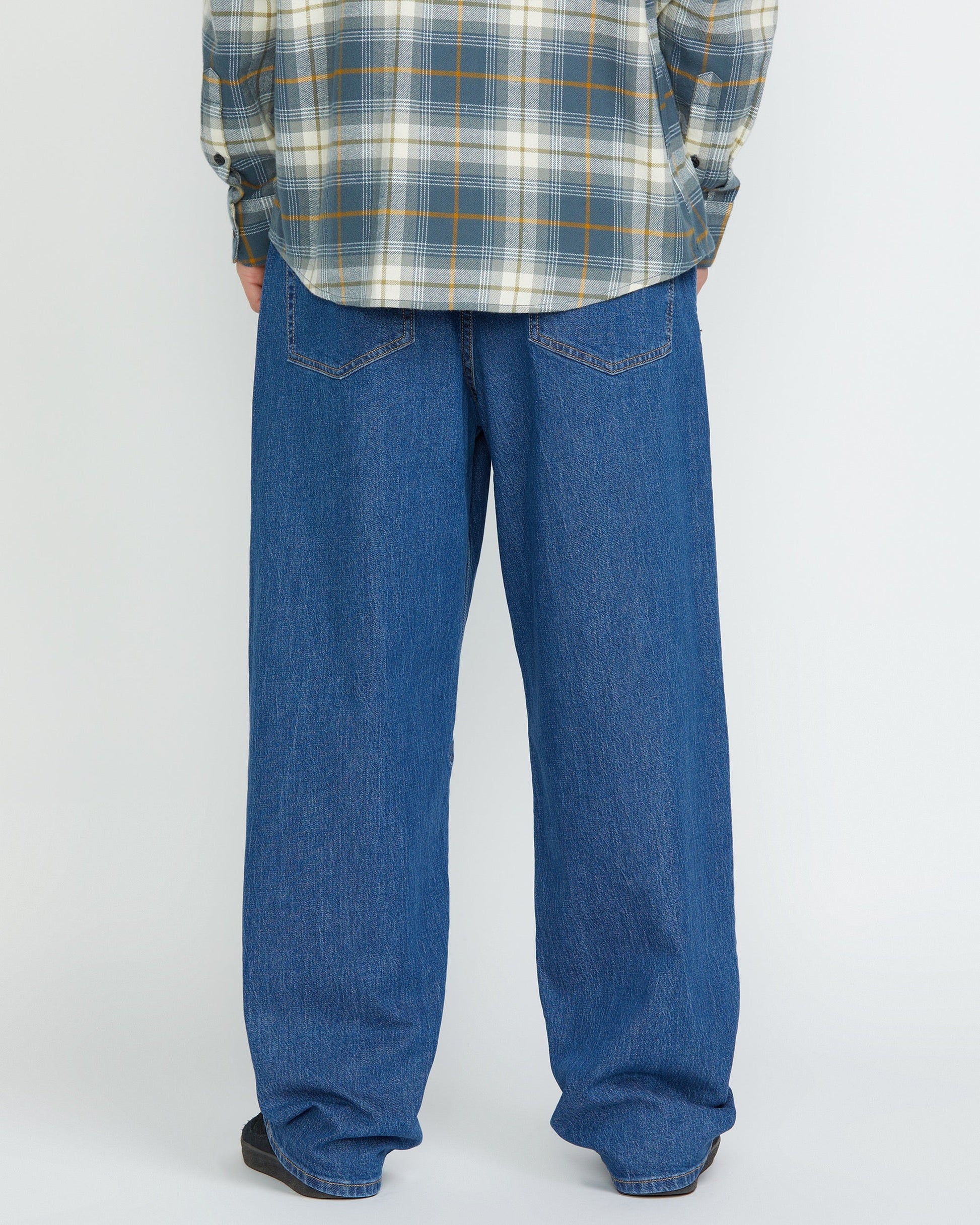 A person stands facing away, showing only the lower half, wearing Volcom Men's Billow Denim Pants and a gray plaid shirt against a plain light background.