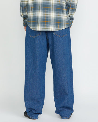 A person stands facing away, showing only the lower half, wearing Volcom Men's Billow Denim Pants and a gray plaid shirt against a plain light background.
