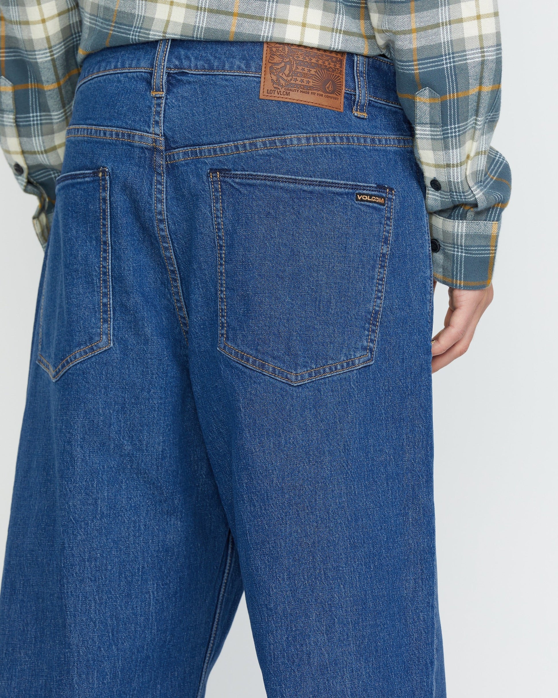 A person wears Volcom Men's Billow Denim Pants, loose fit blue jeans with a brown leather patch on the waistband and a visible Volcom label on the back pocket, paired with a plaid shirt. The image shows the back view of these low-rise jeans.