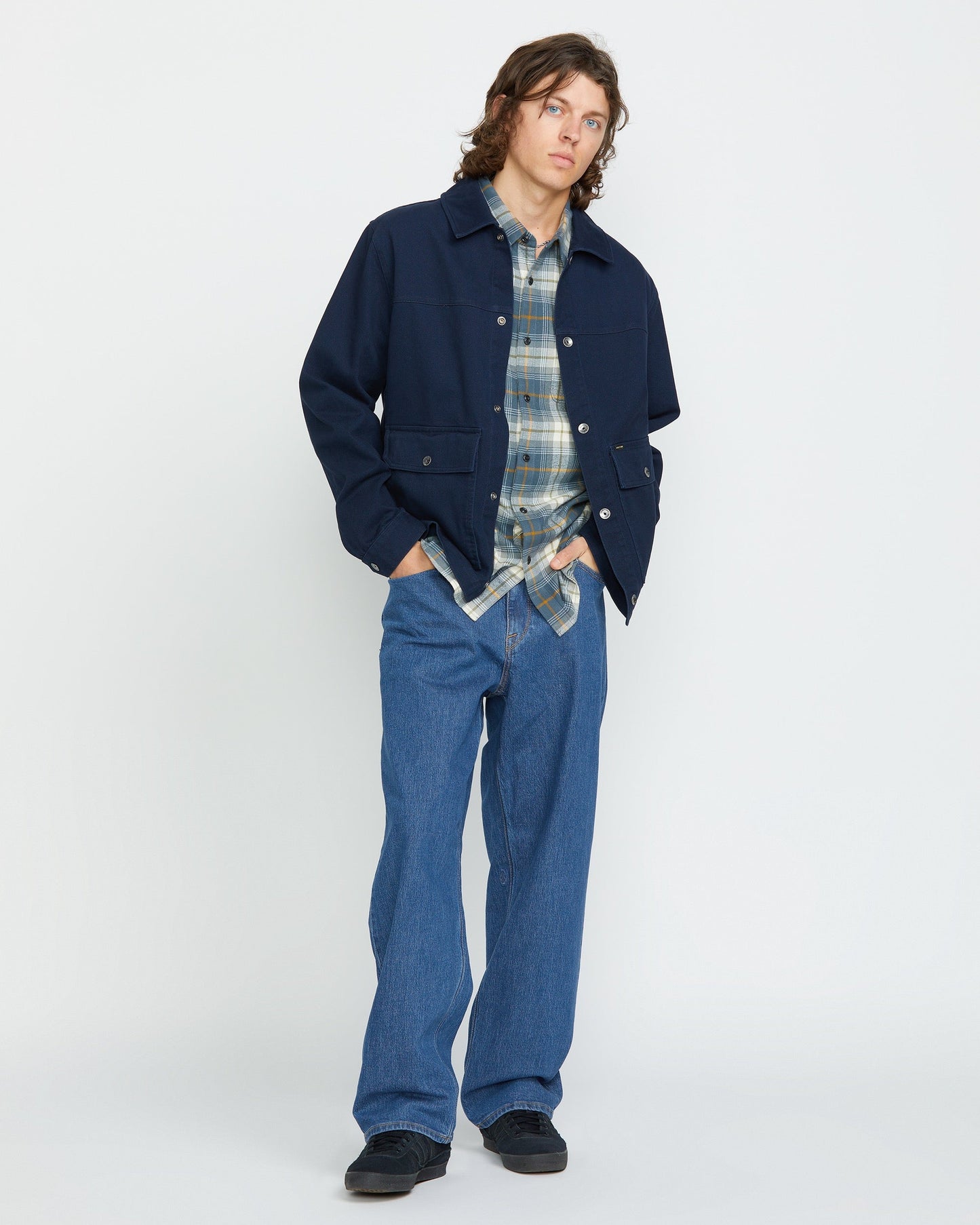 A young man with wavy brown hair stands against a plain white background, wearing a navy jacket over a blue plaid shirt, Volcom Men's Billow Denim Pants in a loose fit, and black shoes. His hands are in his jacket pockets as he looks at the camera.