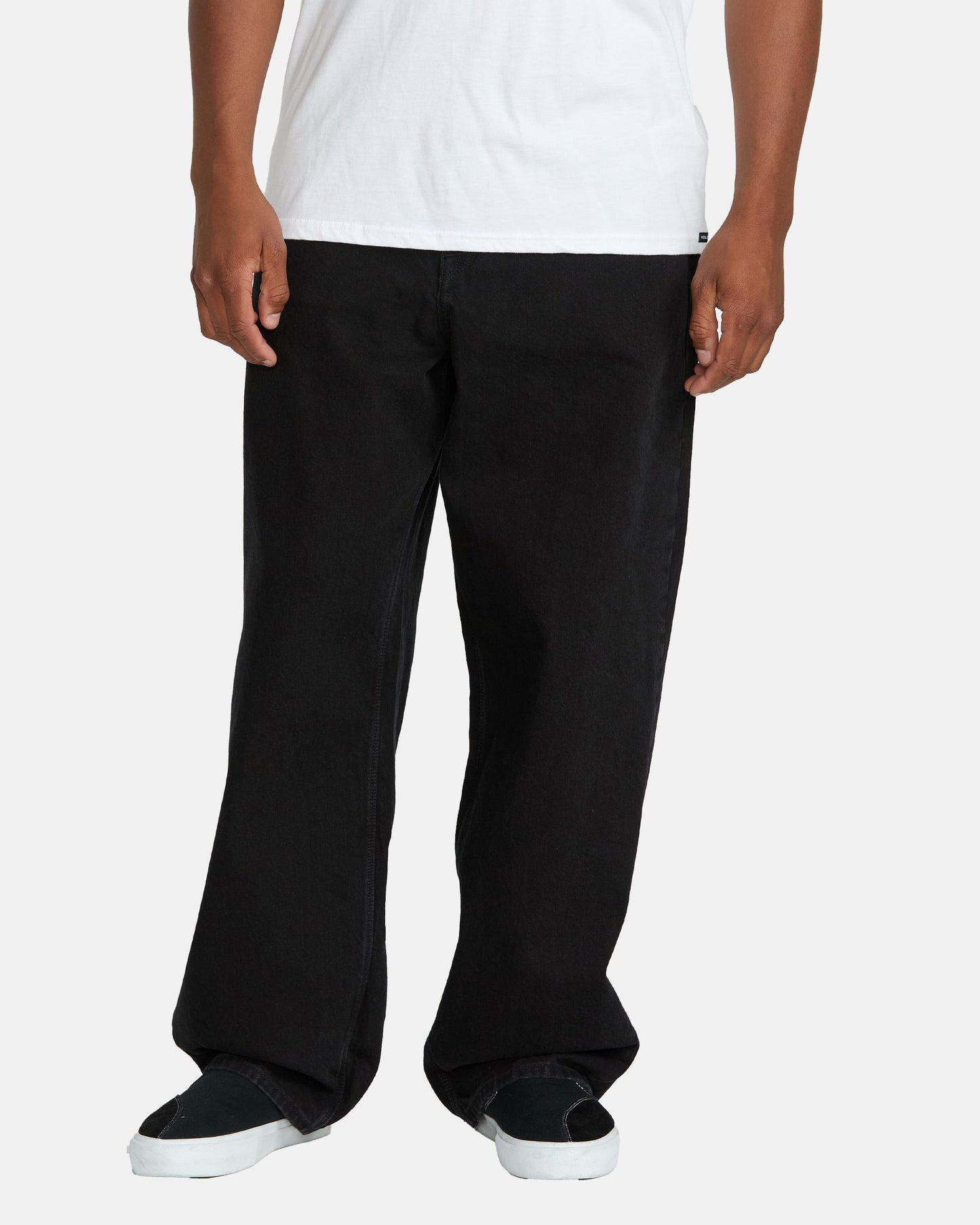 A person wearing Volcom Men's Billow Denim Pants in black, a white t-shirt, and black slip-on sneakers with white soles stands against a plain light background.