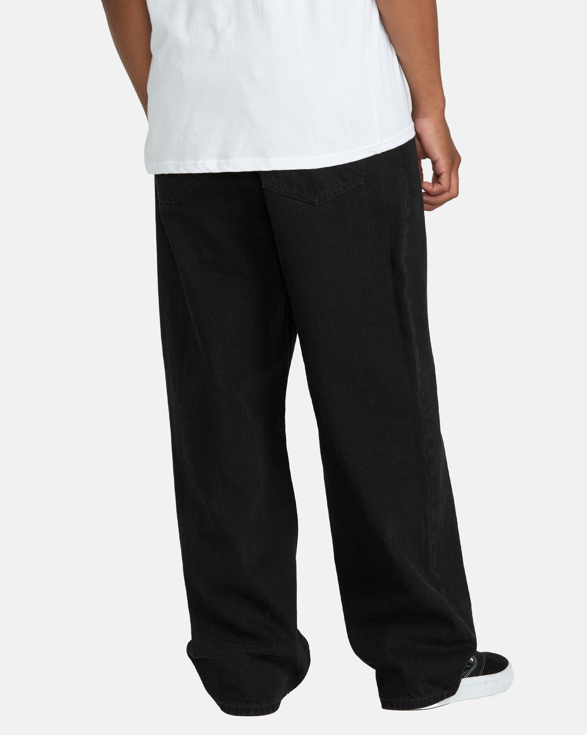A person wearing a white T-shirt, Volcom Men's Billow Denim Pants, and black-and-white sneakers stands facing away from the camera against a plain background.