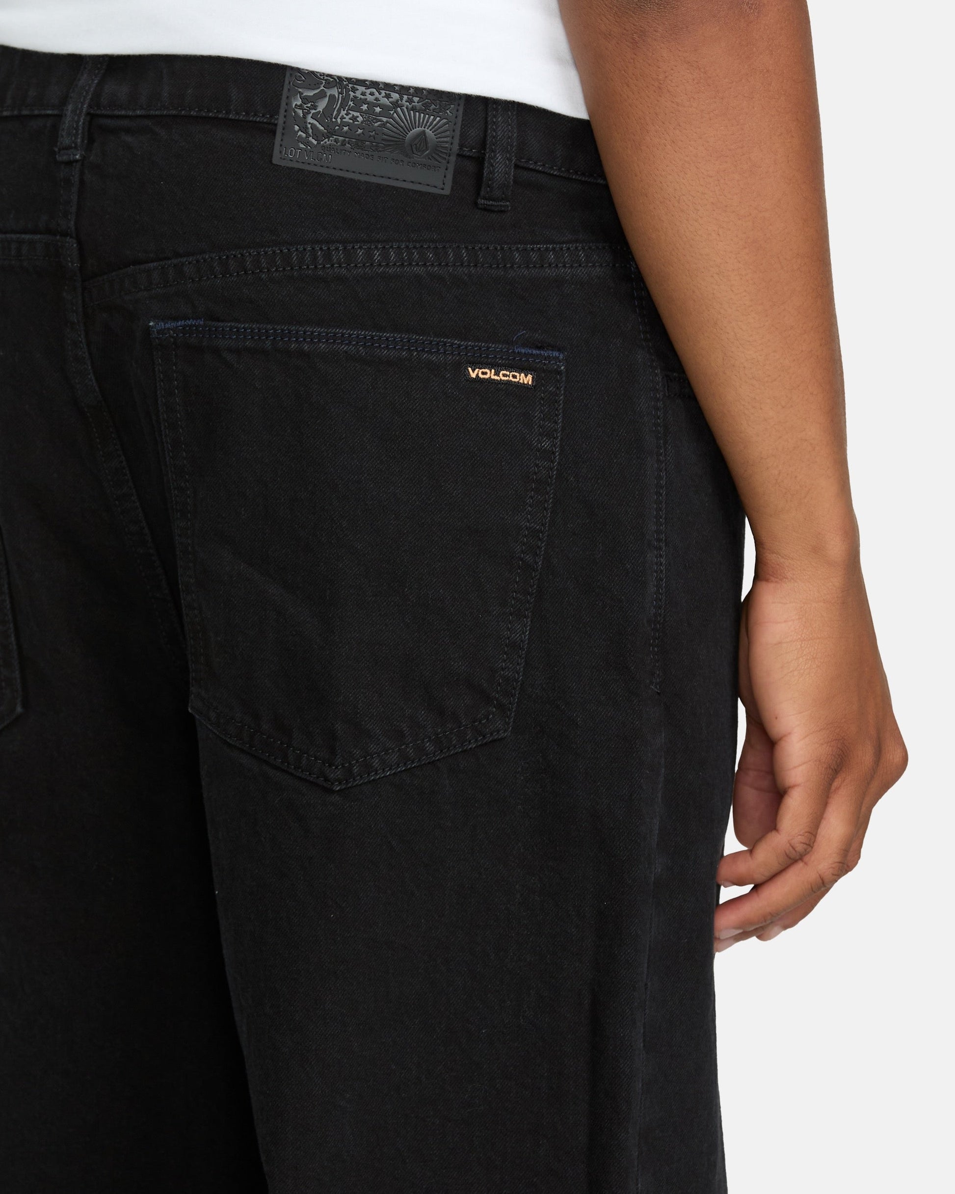 A person wearing loose fit Men's Billow Denim Pants by Volcom, featuring a visible logo on the back pocket; their right hand rests at their side, and part of a white shirt is visible at the waist.