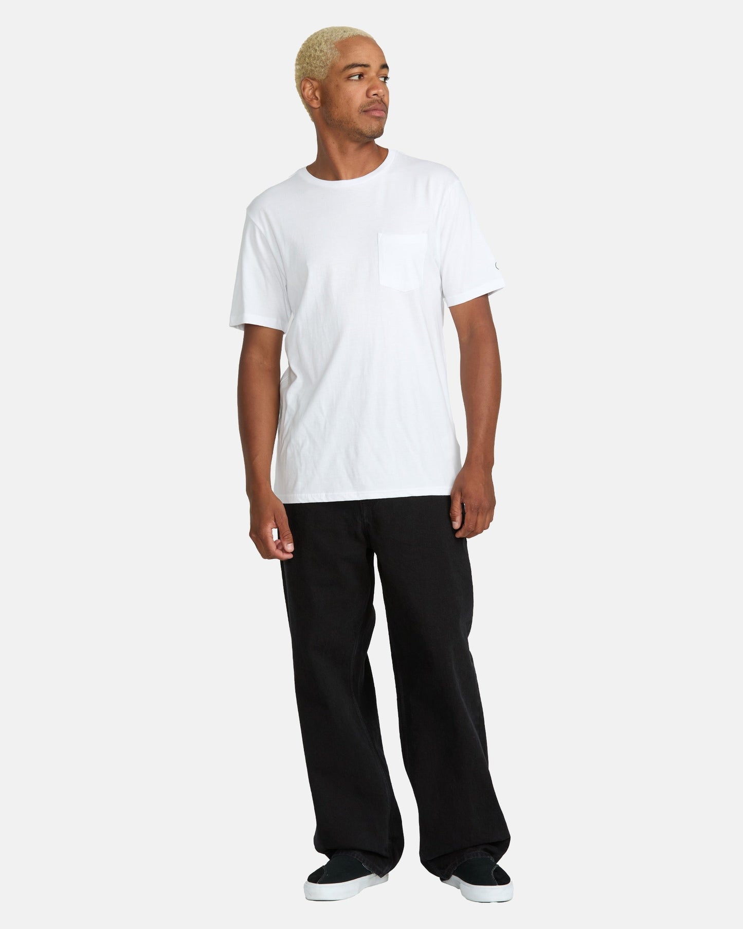 A person with short blonde hair wears a white short-sleeve pocket t-shirt, black Volcom Men's Billow Denim Pants in a loose wide-leg fit, and black shoes, standing against a plain light gray background.