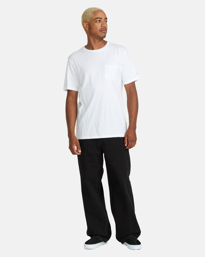 A person with short blonde hair wears a white short-sleeve pocket t-shirt, black Volcom Men's Billow Denim Pants in a loose wide-leg fit, and black shoes, standing against a plain light gray background.