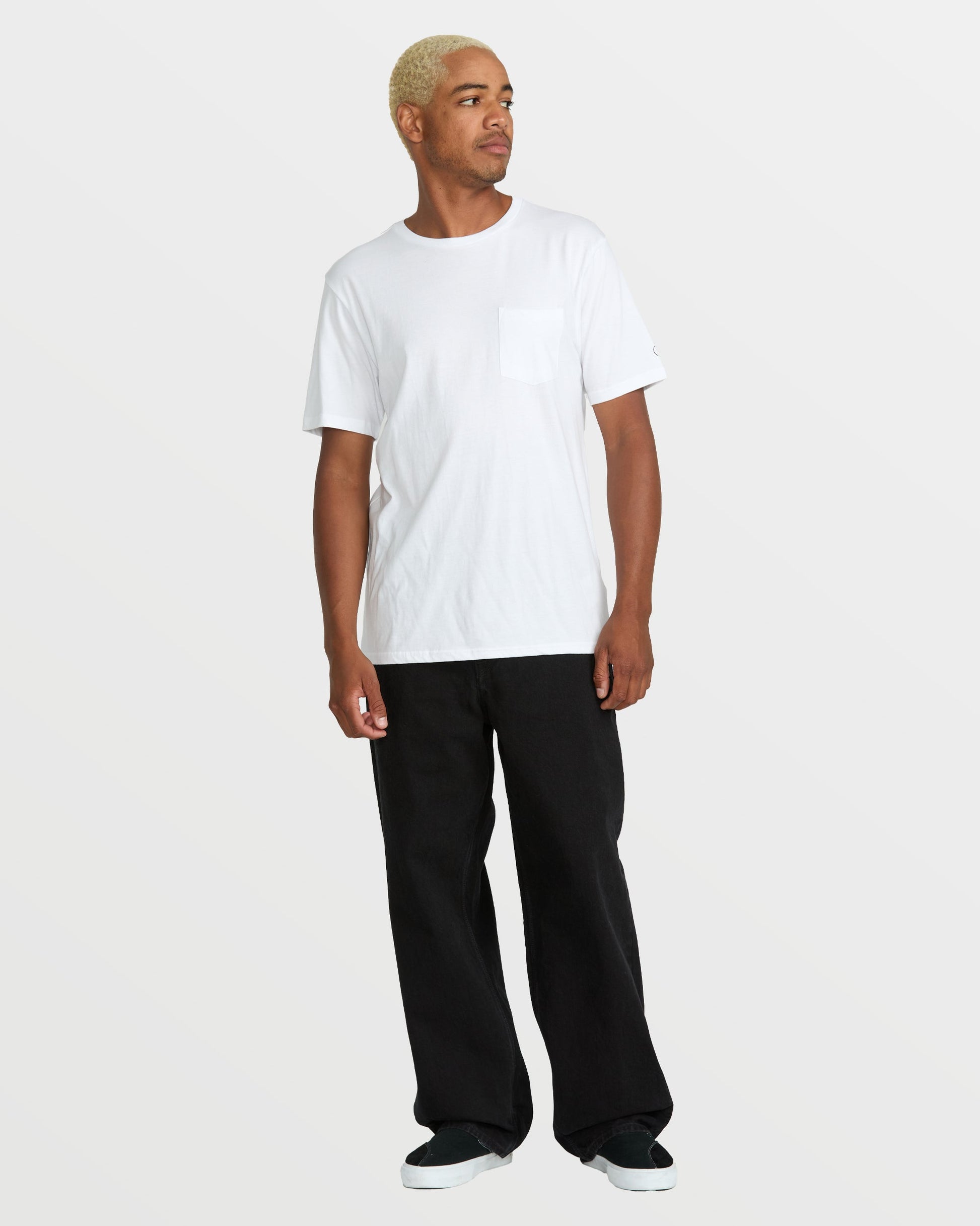 A person with short blond hair wears a white short-sleeve pocket t-shirt, Volcom Men's Billow Denim jeans in a loose fit, and black slip-on shoes, standing against a plain light gray background and looking to the side.