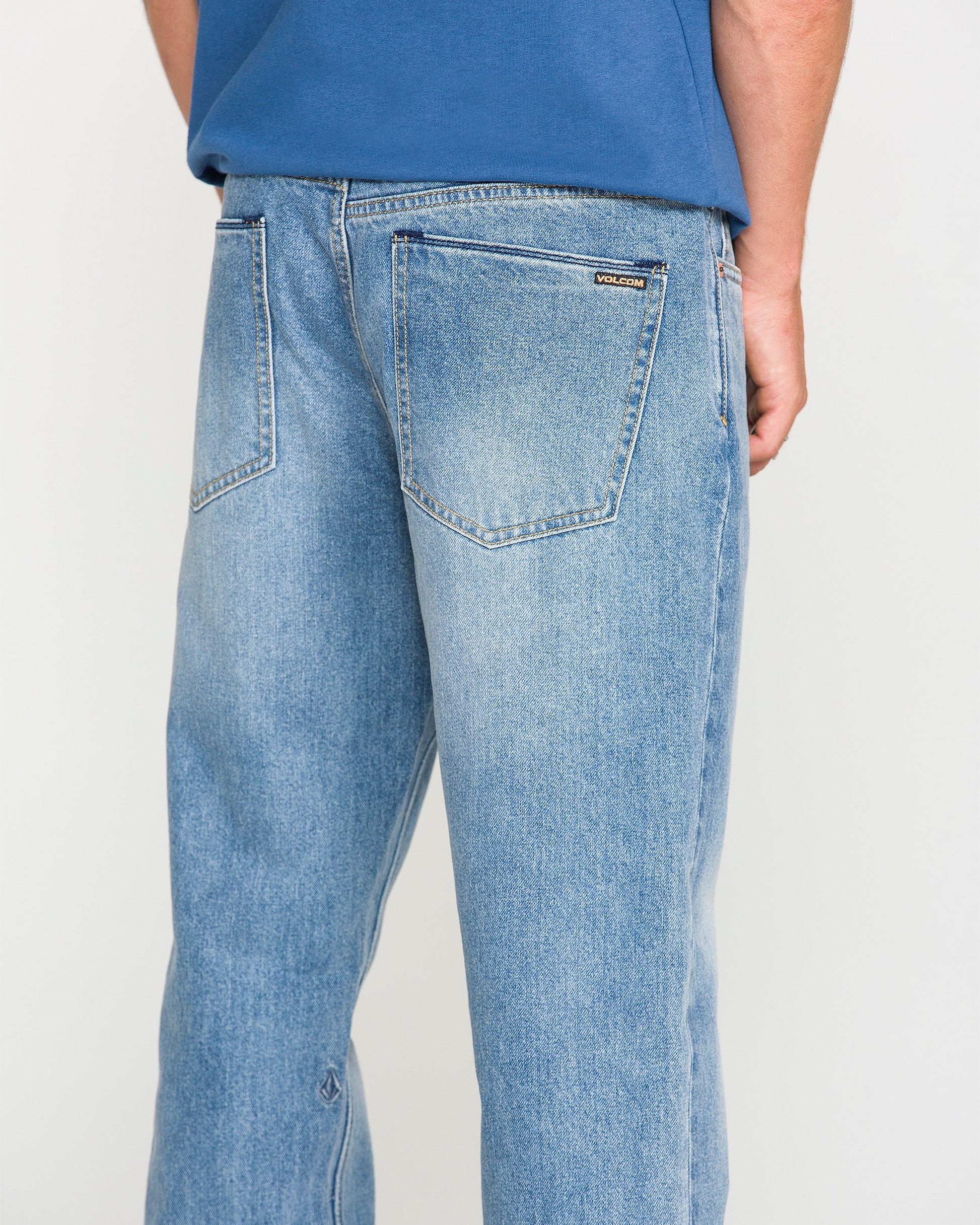 A person wearing Volcom Men's Solver Denim Pants in Freedom Blue and a blue shirt, shown from the back. The stretch denim jeans feature two back pockets and a small Volcom label on the right pocket.