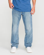 A person wearing a Freedom Blue T-shirt, Volcom Men's Solver Denim Pants in light blue stretch denim with a modern straight leg cut, and white sneakers stands facing forward against a plain white background. Their head is not visible in the image.