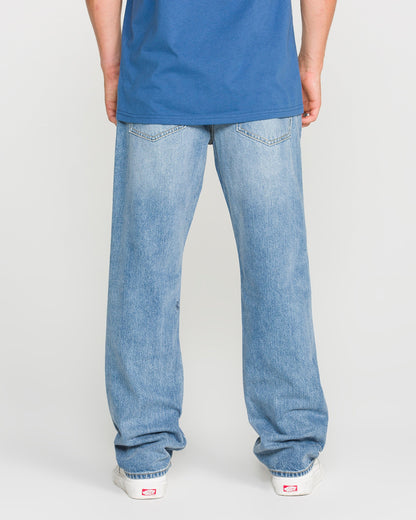 A person stands facing away, wearing a blue t-shirt, white sneakers, and Volcom Men's Solver Denim Pants in Freedom Blue—modern straight-leg jeans in stretchy light blue denim. The background is plain and light-colored.