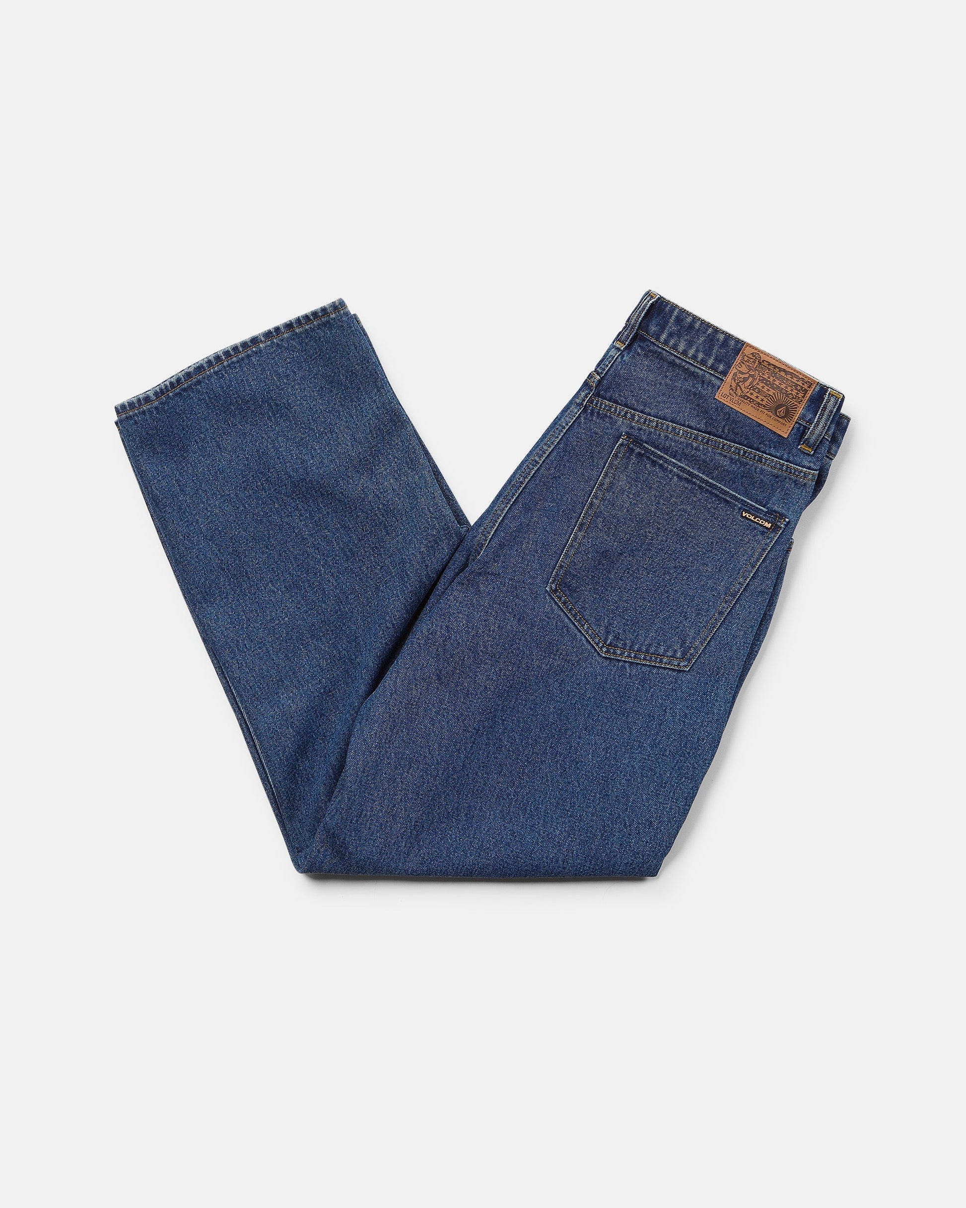 A pair of Volcom Men's Billow Denim Pants in dark indigo is neatly folded, showcasing a brown leather patch on the waistband and a visible back pocket. The low rise jeans are displayed against a plain white background.