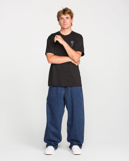 A young man stands against a plain background, wearing a black t-shirt, white sneakers, and Volcom Men's Billow Denim Pants in a loose fit dark indigo wash. One hand is raised near his face as he maintains a neutral expression.
