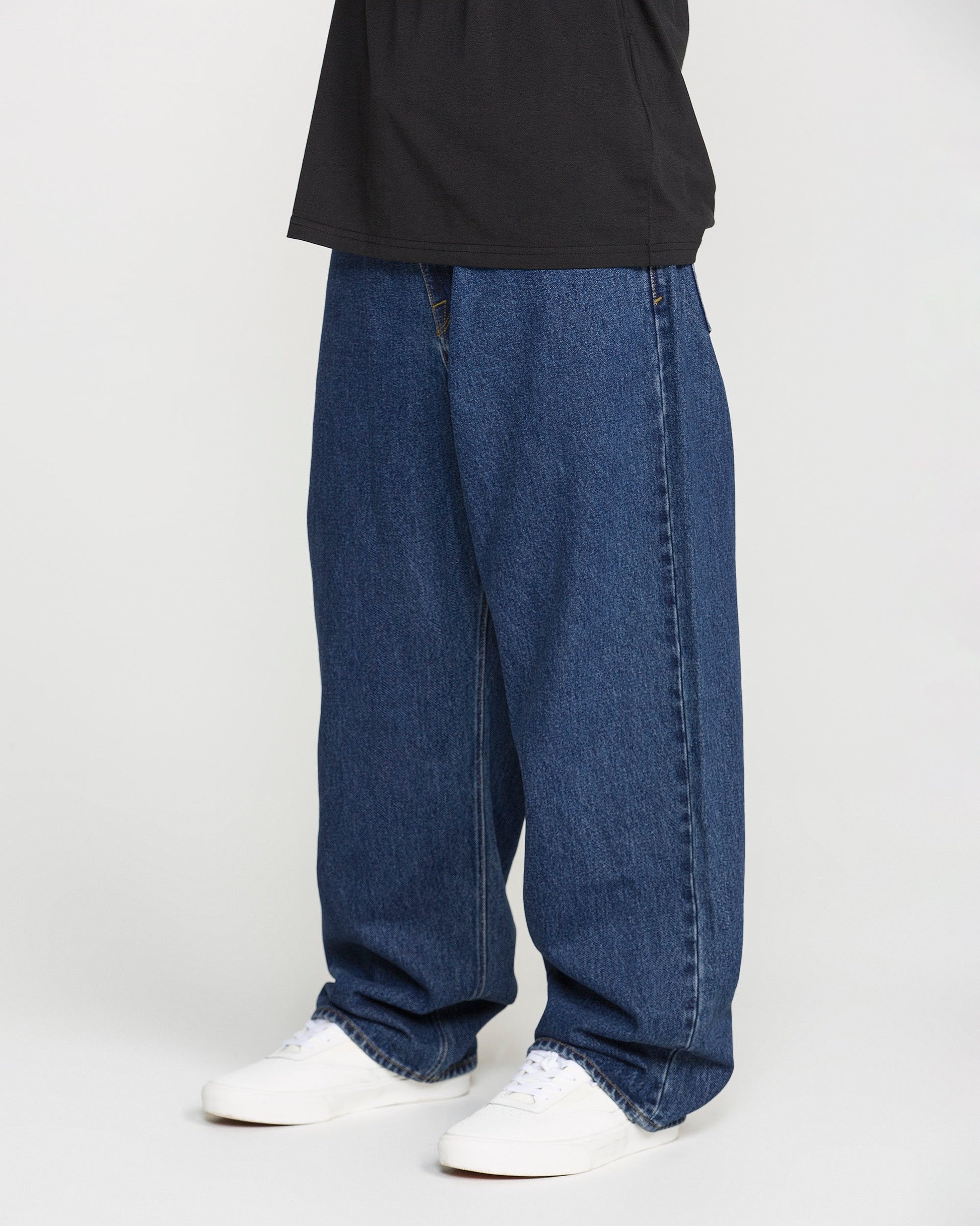 A person wears the Volcom Men's Billow Denim Pants—a loose-fitting, low-rise style in classic blue wash—paired with white sneakers and a black shirt. The image is cropped above the shoulders, highlighting the lower half of the outfit against a plain background.