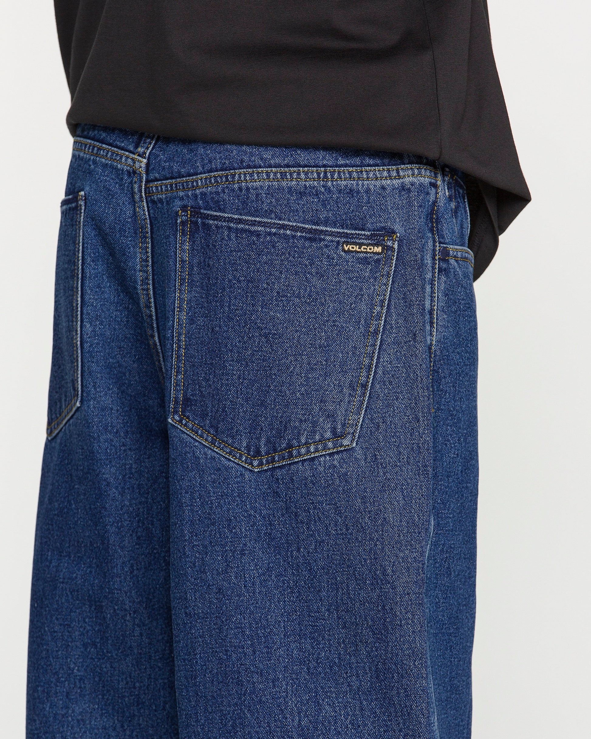 A person is wearing loose fit blue Men's Billow Denim Pants by Volcom and a black shirt, shown from the waist down and from the back, with a focus on the back pocket featuring a small Volcom label. The low rise design of these Volcom jeans adds a relaxed edge to this casual look.