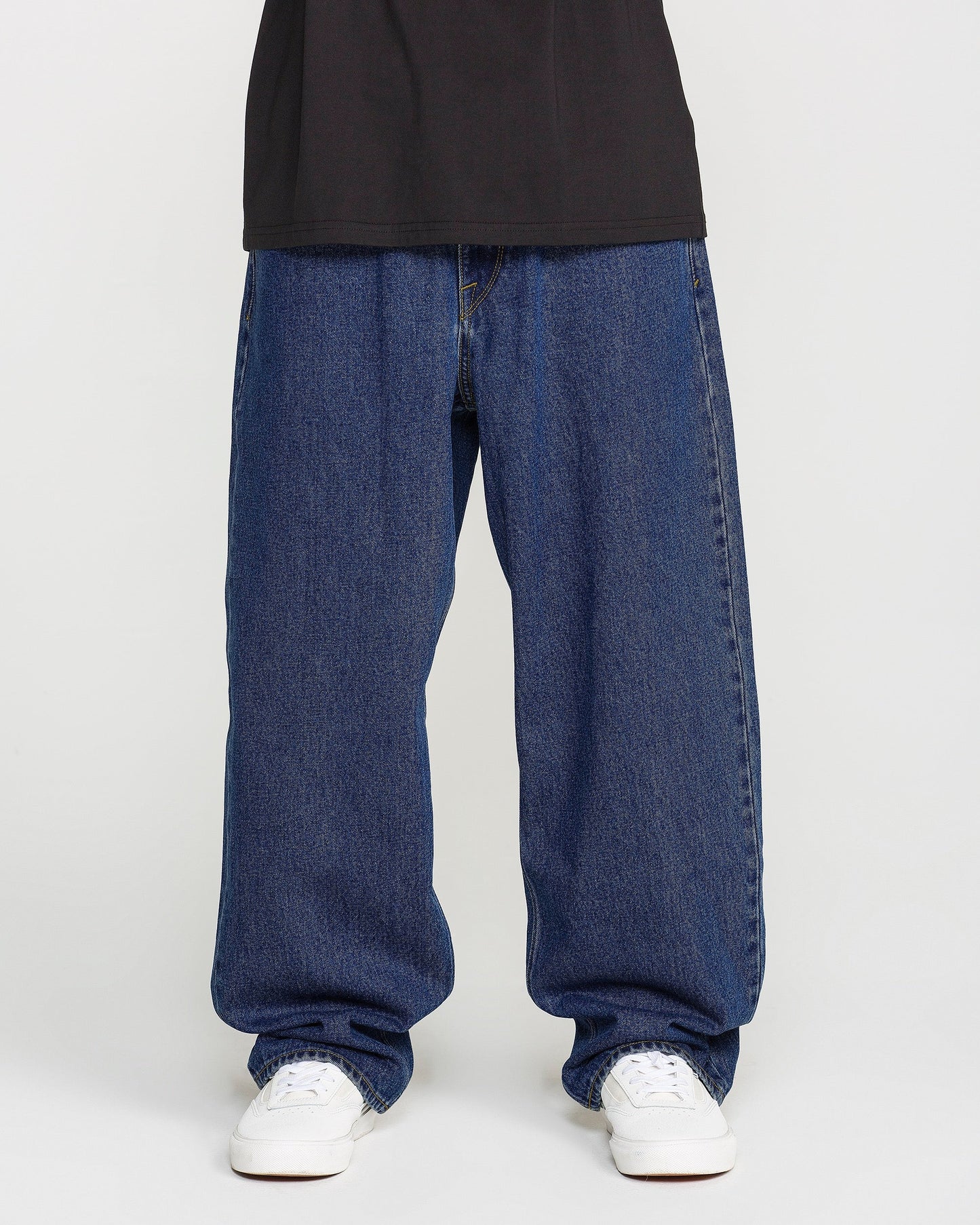 A person is shown from the waist down, wearing Volcom Men's Billow Denim Pants in a loose fit dark indigo wash, paired with a black shirt and white sneakers, standing against a plain white background.