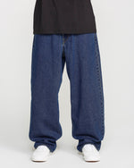 A person is shown from the waist down, wearing Volcom Men's Billow Denim Pants in a loose fit dark indigo wash, paired with a black shirt and white sneakers, standing against a plain white background.
