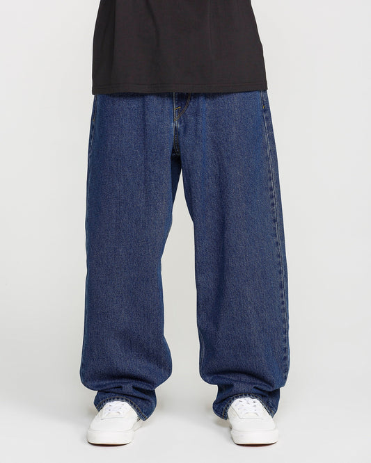 A person is shown from the waist down, wearing Volcom Men's Billow Denim Pants in a loose fit dark indigo wash, paired with a black shirt and white sneakers, standing against a plain white background.