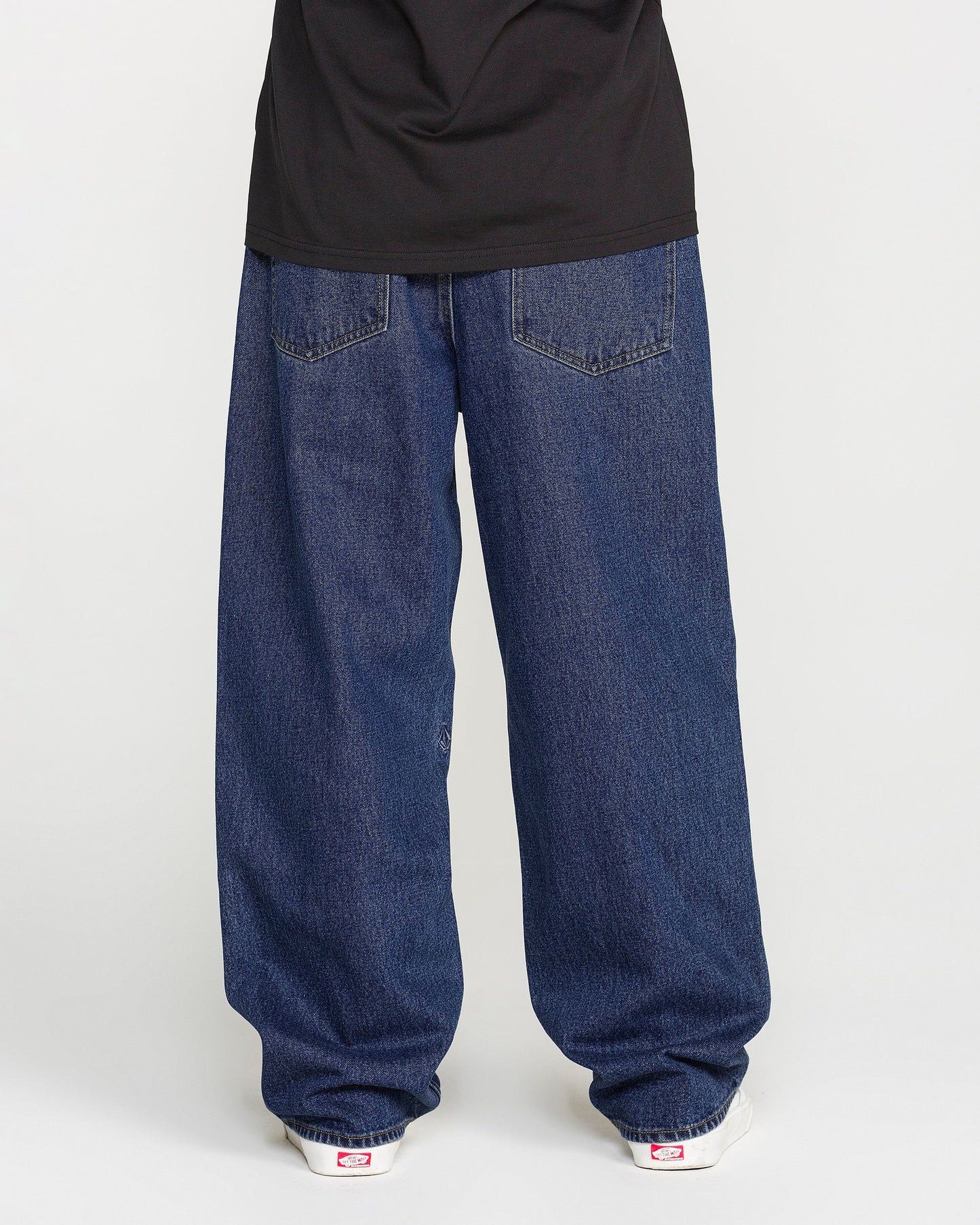 A person is standing against a plain light background, photographed from behind to show only the lower half of their body. They are wearing Volcom Men's Billow Denim Pants in a dark indigo wash, paired with a black shirt and white sneakers.