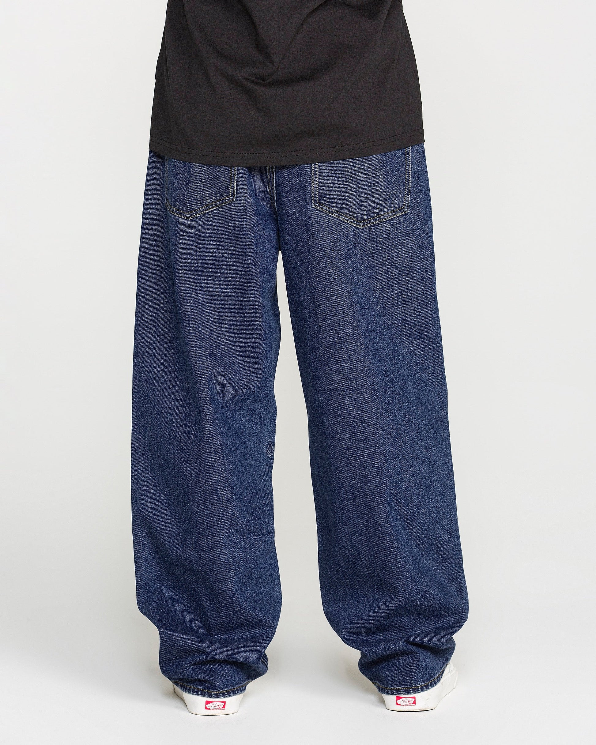 A person is standing against a plain light background, photographed from behind to show only the lower half of their body. They are wearing Volcom Men's Billow Denim Pants in a dark indigo wash, paired with a black shirt and white sneakers.