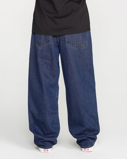 A person is standing against a plain light background, photographed from behind to show only the lower half of their body. They are wearing Volcom Men's Billow Denim Pants in a dark indigo wash, paired with a black shirt and white sneakers.