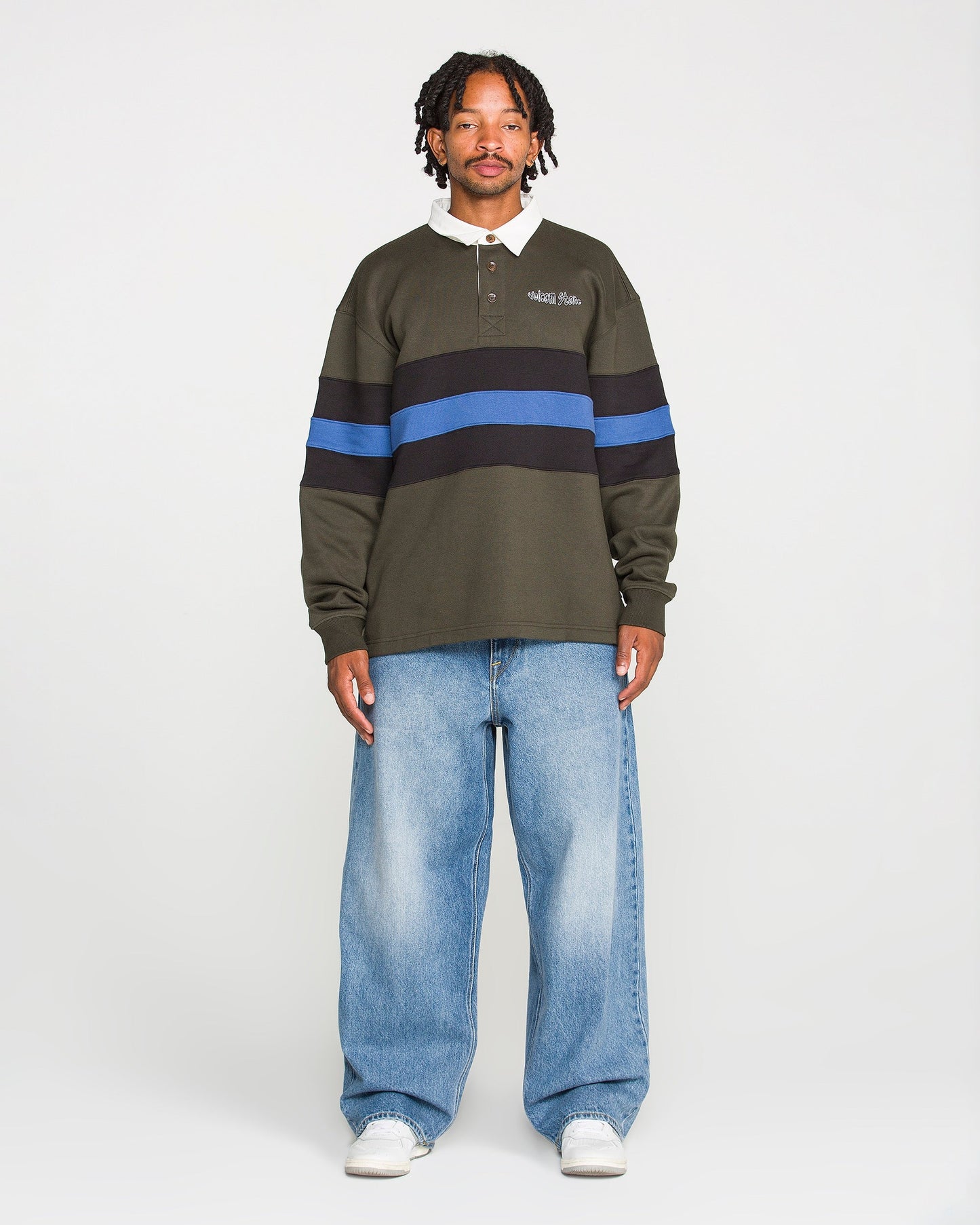 A man stands facing forward, wearing a dark green rugby shirt with blue and black horizontal stripes, Volcom Men's Chillow Denim Pants, and white sneakers against a plain background.