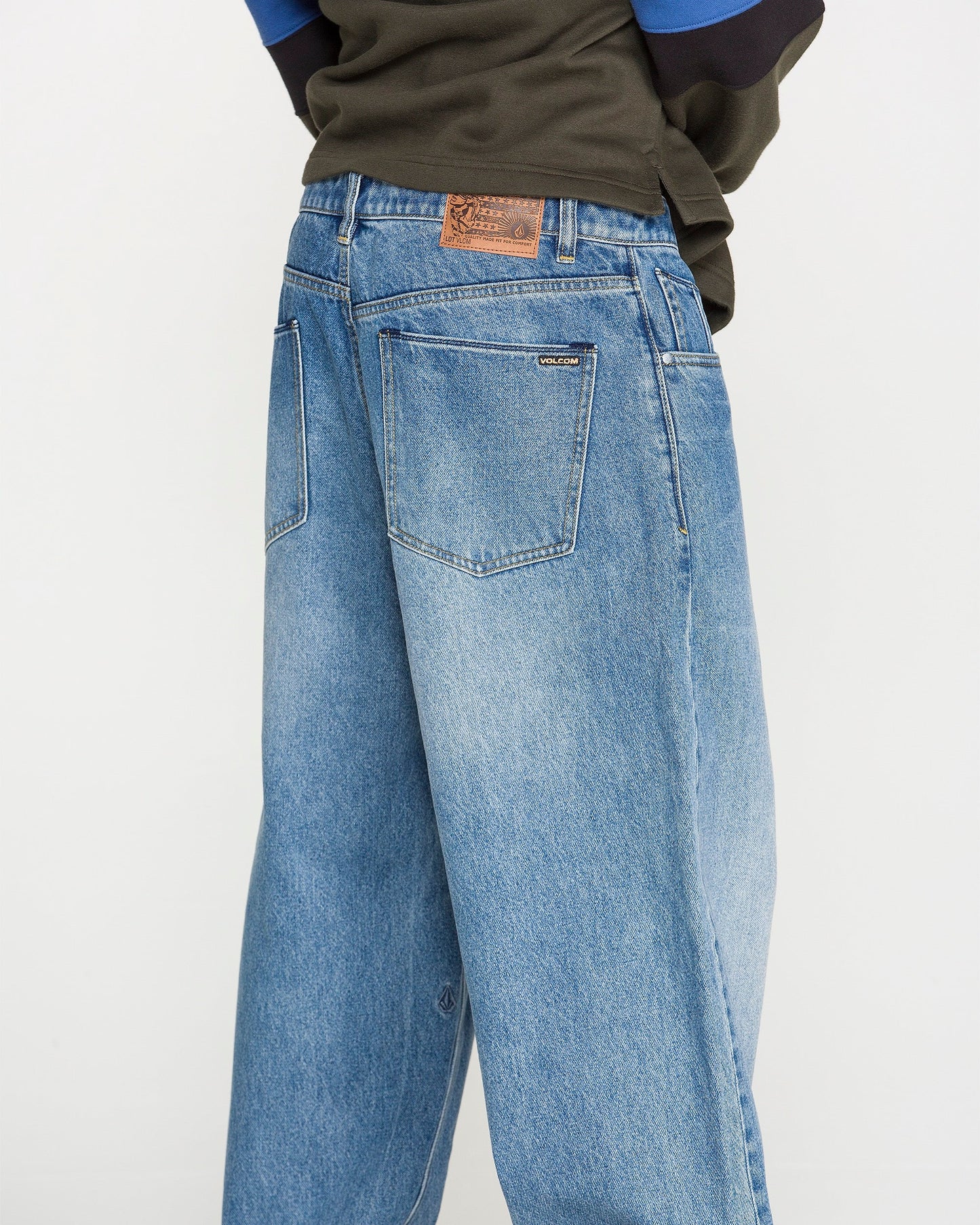 A person wearing Volcom Men's Chillow Denim Pants, featuring a relaxed fit in blue denim with visible back pockets and a brown patch on the waistband, is photographed from behind against a plain background.