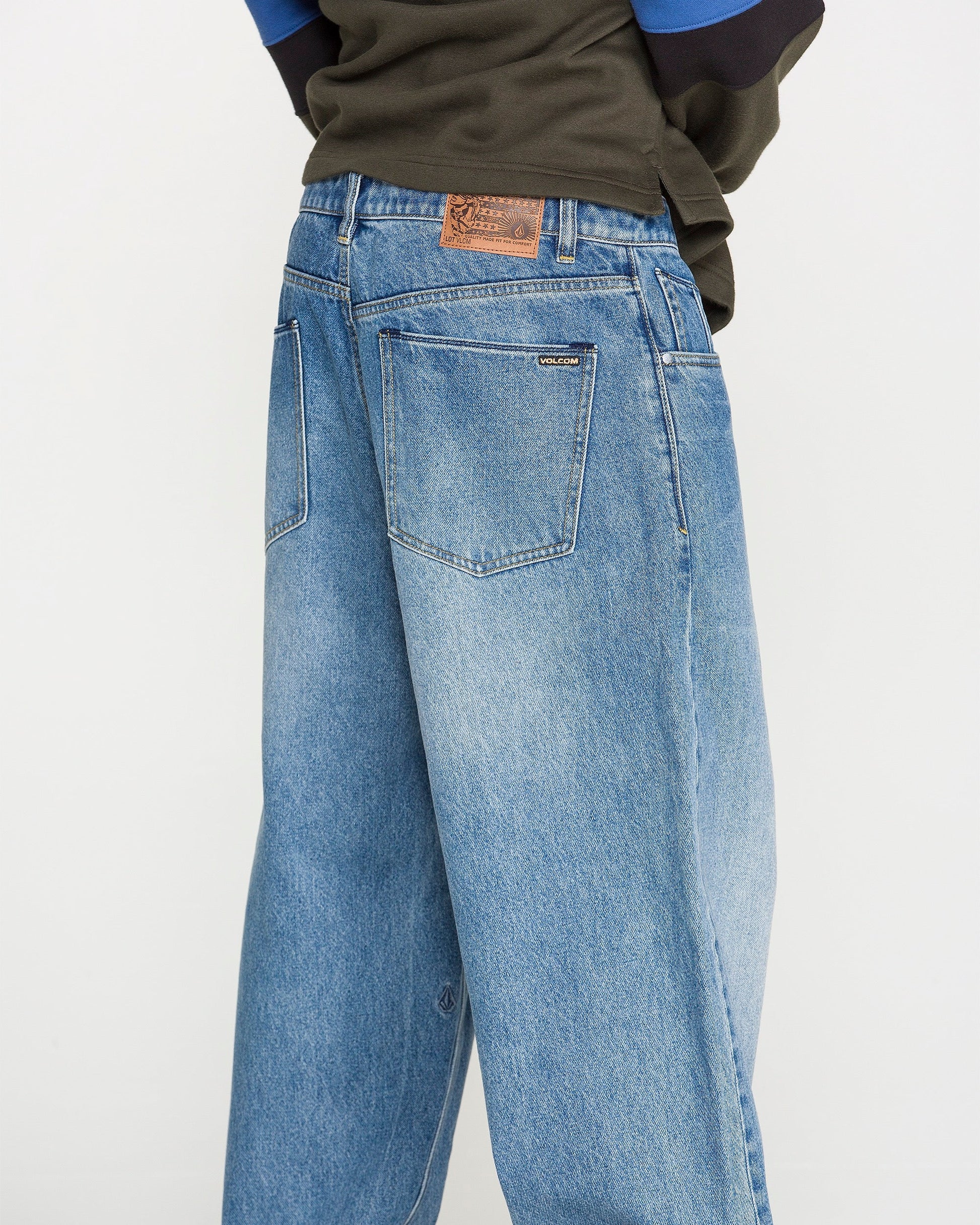 A person wearing Volcom Men's Chillow Denim Pants, featuring a relaxed fit in blue denim with visible back pockets and a brown patch on the waistband, is photographed from behind against a plain background.