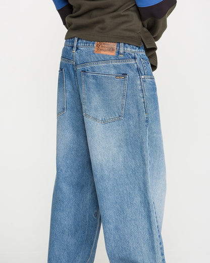 A person wearing Volcom Men's Chillow Denim Pants, featuring a relaxed fit in blue denim with visible back pockets and a brown patch on the waistband, is photographed from behind against a plain background.