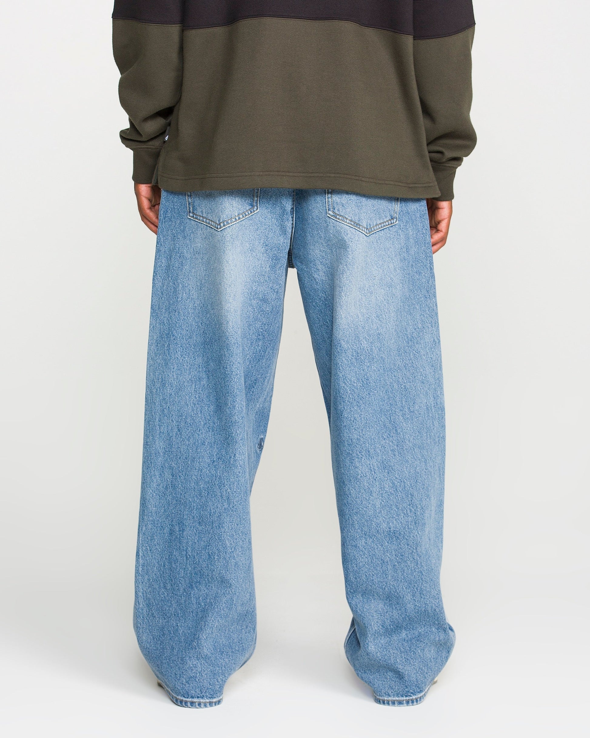A person stands with their back to the camera against a plain white background, wearing a dark olive green sweatshirt and Volcom Men's Chillow Denim Pants in a loose-fitting, Euro Light Blue wash.