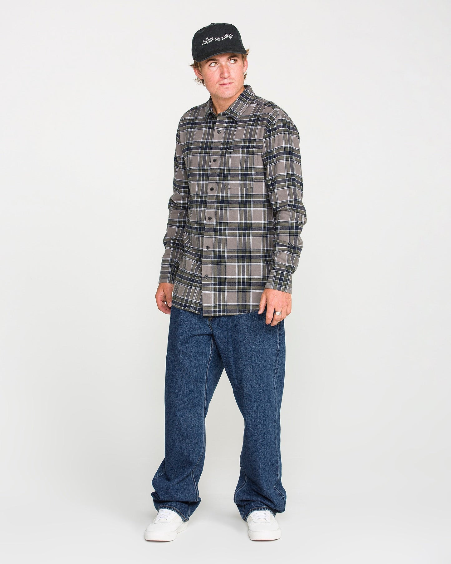 A man wearing a black cap, plaid button-up shirt, Volcom Men's Pipe Dreams Denim Pants in dark indigo, and white sneakers stands against a plain white background, looking slightly to his left.