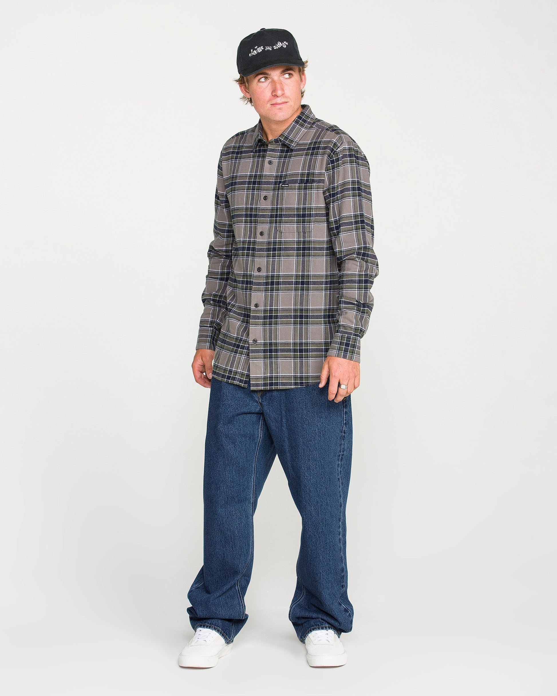 A man wearing a black cap, plaid button-up shirt, Volcom Men's Pipe Dreams Denim Pants in dark indigo, and white sneakers stands against a plain white background, looking slightly to his left.