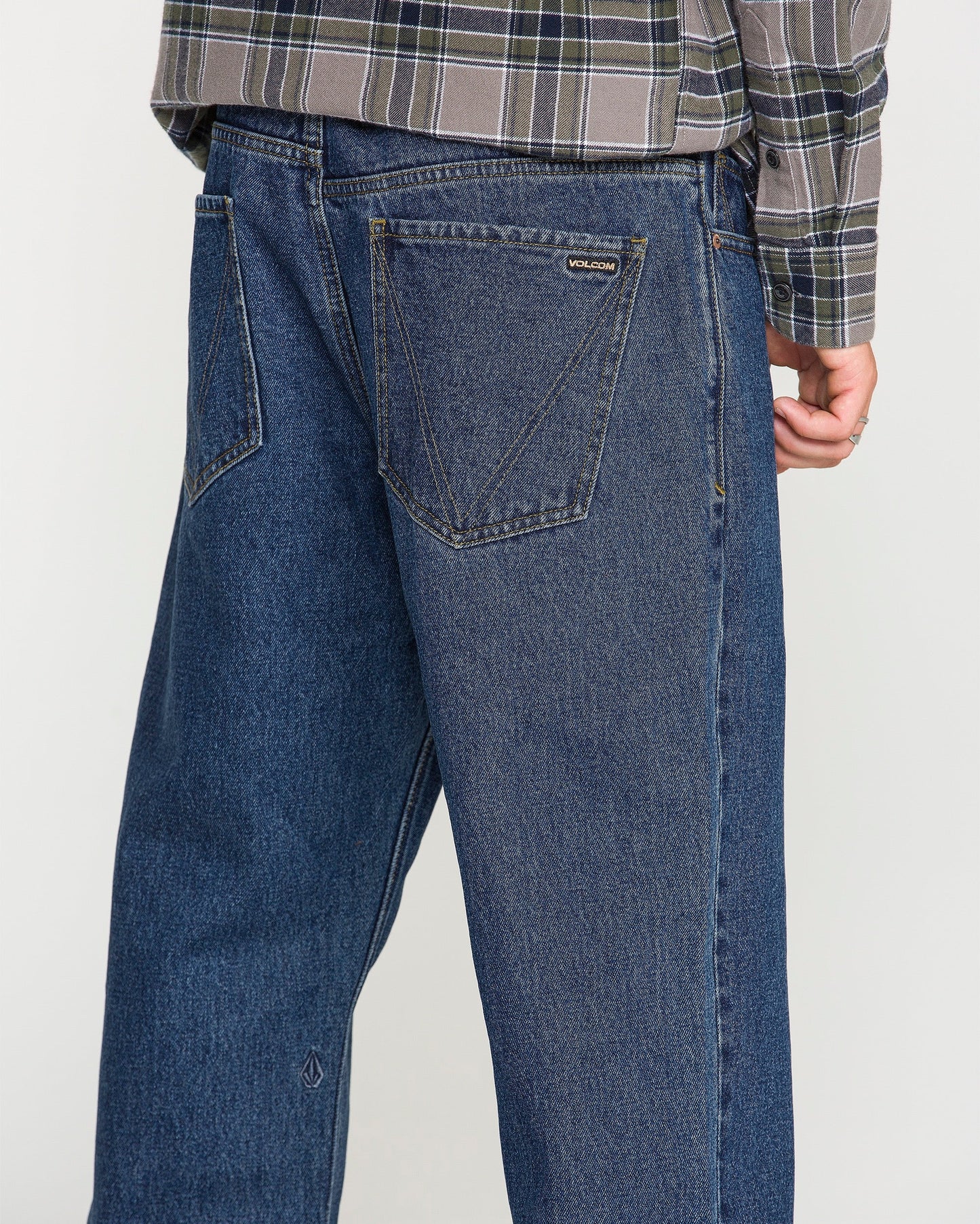 A person is shown from the back wearing Volcom Men's Pipe Dreams Denim Pants in a relaxed straight fit dark indigo denim, paired with a gray plaid shirt. One hand is partially visible and the jeans feature large back pockets and a small Volcom brand tag.