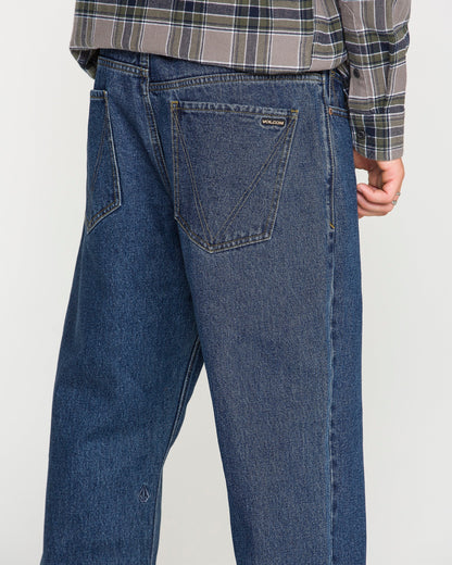 A person is shown from the back wearing Volcom Men's Pipe Dreams Denim Pants in a relaxed straight fit dark indigo denim, paired with a gray plaid shirt. One hand is partially visible and the jeans feature large back pockets and a small Volcom brand tag.