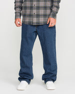 A person standing against a plain background, shown from the shoulders down, is wearing a gray plaid shirt, white sneakers, and relaxed straight fit Men's Pipe Dreams Denim Pants by Volcom in dark indigo denim.