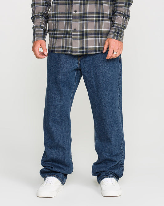 A person standing against a plain background, shown from the shoulders down, is wearing a gray plaid shirt, white sneakers, and relaxed straight fit Men's Pipe Dreams Denim Pants by Volcom in dark indigo denim.